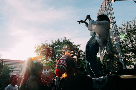 A performer is on stage energetically engaging with the crowd, while audience members cheer and raise their hands. The backlighting creates a silhouette effect around the performer. Trees and a building are visible in the background, with sunlight streaming through the scene.