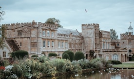 A historic stone mansion with intricate architectural details, including arched windows and battlements. Manicured gardens with colorful flowers and trimmed hedges surround the building. A pond in the foreground reflects part of the structure and vegetation. The scene is under a cloudy sky, adding to the serene atmosphere.
