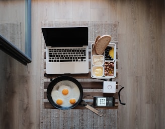 A neatly arranged setup on a wooden table featuring a laptop, a plate of fried eggs in a pan, a smartphone playing music, and a meal prep container with assorted food items including bread, butter, blueberries, and olives.