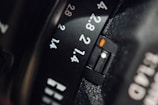 A close-up view of a camera lens showing aperture settings marked with numbers like 1.4, 2, 2.8, and 4. The black surface features engraved white numbers, with a small orange marker highlighting the aperture setting.