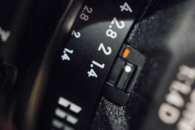 A close-up view of a camera lens showing aperture settings marked with numbers like 1.4, 2, 2.8, and 4. The black surface features engraved white numbers, with a small orange marker highlighting the aperture setting.