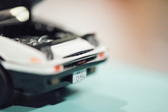 A close-up of a toy car with its hood open, revealing parts of the engine interior. The focus is on the front of the car, featuring a visible license plate with Japanese characters.