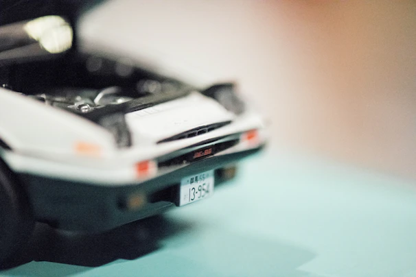 A close-up of a toy car with its hood open, revealing parts of the engine interior. The focus is on the front of the car, featuring a visible license plate with Japanese characters.