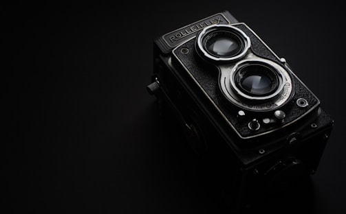 A vintage Rolleiflex camera with dual lenses is set against a dark background. The camera features distinct metallic and textured elements, with prominent glass lenses reflecting light. The overall design portrays a sense of classic elegance and craftsmanship.