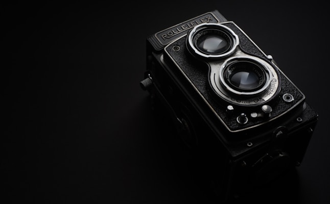 A vintage Rolleiflex camera with dual lenses is set against a dark background. The camera features distinct metallic and textured elements, with prominent glass lenses reflecting light. The overall design portrays a sense of classic elegance and craftsmanship.