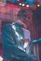 A young person wearing round glasses and a denim jacket stands illuminated by string lights. The background is softly blurred with colorful bokeh effects, adding a dreamy atmosphere. The expression is contemplative, with soft light highlighting facial features.