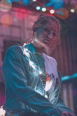 A young person wearing round glasses and a denim jacket stands illuminated by string lights. The background is softly blurred with colorful bokeh effects, adding a dreamy atmosphere. The expression is contemplative, with soft light highlighting facial features.