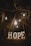 In a dimly lit forest setting, several glowing light bulbs hang from tree branches. Below them, illuminated letters spell out the word 'HOPE'. The scene is surrounded by dark trees and leaves, creating a mysterious atmosphere.