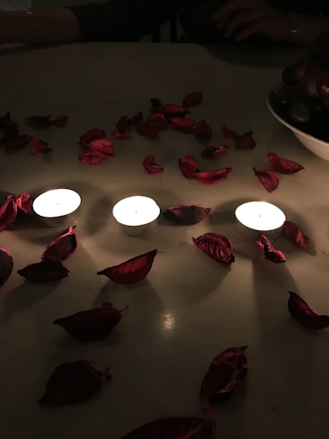 Four lit tealight candles are arranged on a surface surrounded by scattered red rose petals. The lighting is dim, creating a cozy and intimate atmosphere. A bowl of dark grapes is partially visible on the right side of the image.