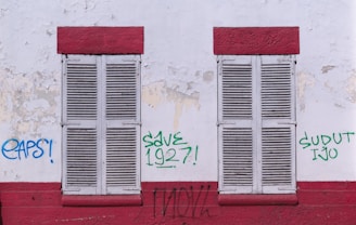 Two closed wooden shutters, painted white, are set within a weathered wall with peeling paint. The wall is marked with graffiti in various colors, including blue and green, with phrases such as 'EAPS!', 'SAVE 1927!', and 'SUDUT IJO'. Red horizontal bands frame the top and bottom of the shutters, adding contrast to the otherwise muted and worn surface.