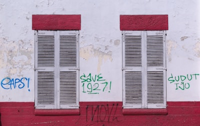 Two closed wooden shutters, painted white, are set within a weathered wall with peeling paint. The wall is marked with graffiti in various colors, including blue and green, with phrases such as 'EAPS!', 'SAVE 1927!', and 'SUDUT IJO'. Red horizontal bands frame the top and bottom of the shutters, adding contrast to the otherwise muted and worn surface.