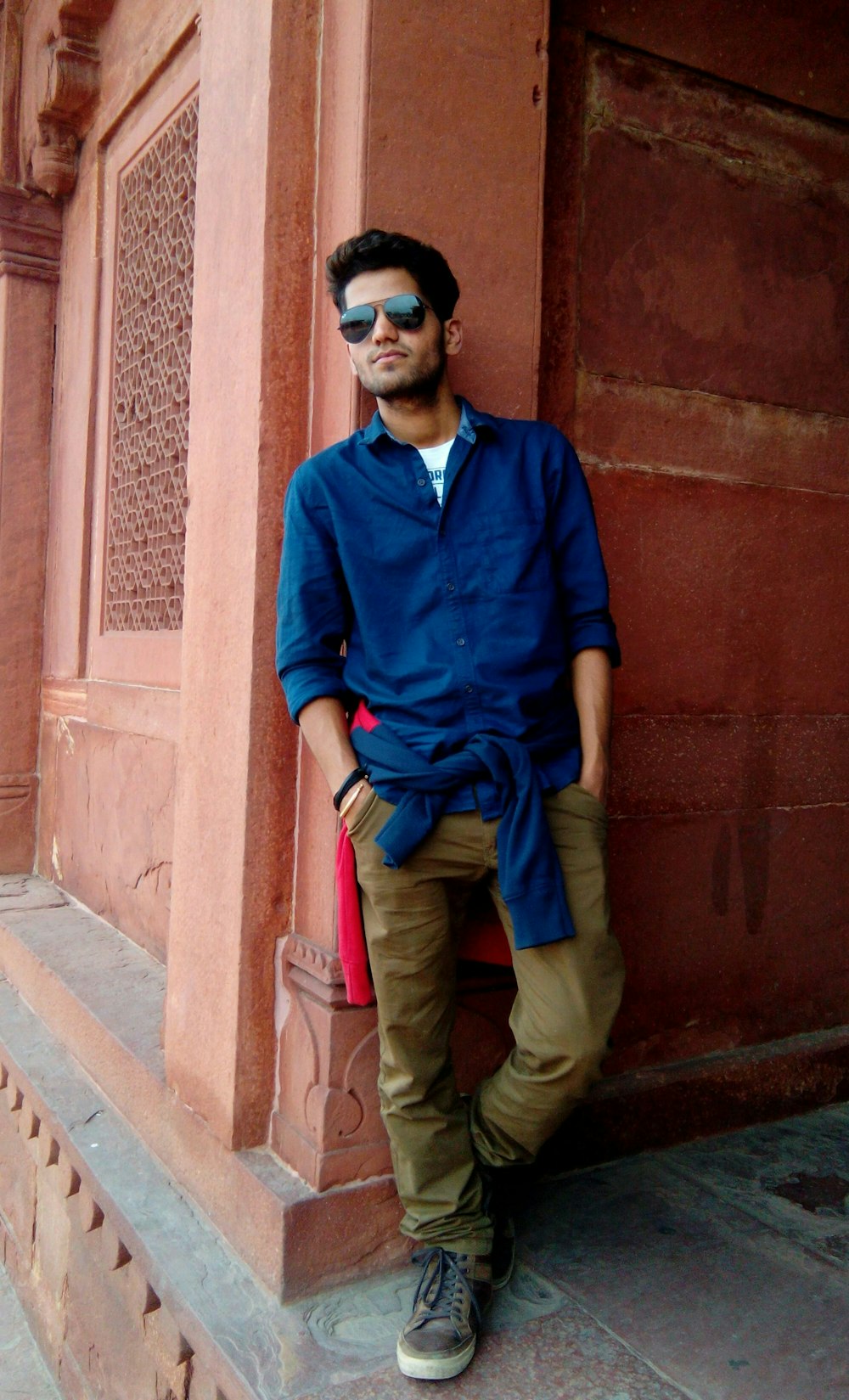 man leaning on brown concrete post wearing blue dress shirt and brown pants