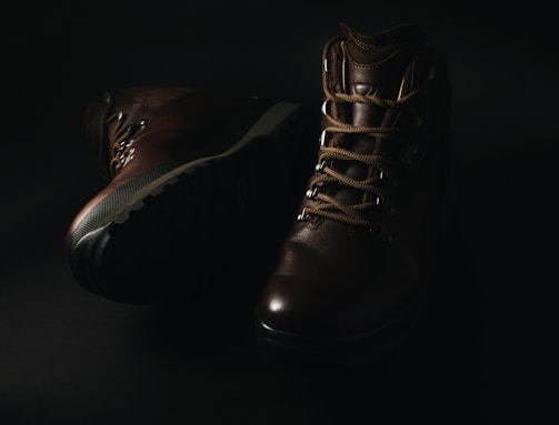A pair of dark brown leather hiking boots with thick soles and sturdy laces is displayed against a dark background, with soft lighting highlighting their texture and detail.