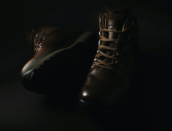 A pair of dark brown leather hiking boots with thick soles and sturdy laces is displayed against a dark background, with soft lighting highlighting their texture and detail.
