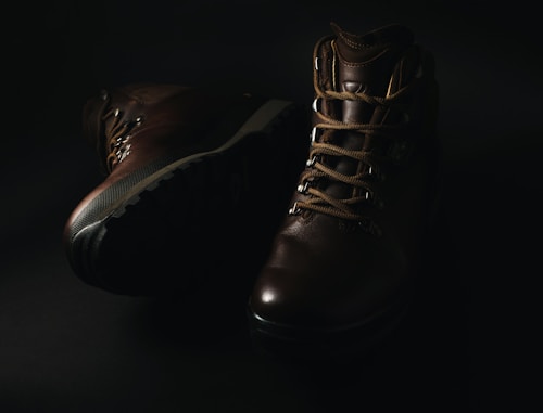 A pair of dark brown leather hiking boots with thick soles and sturdy laces is displayed against a dark background, with soft lighting highlighting their texture and detail.