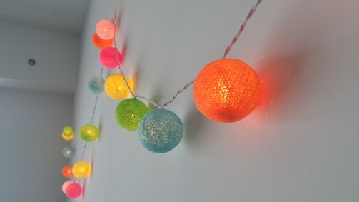 A string of colorful spherical lights hangs along a white wall. The bulbs are illuminated, showcasing a variety of colors including orange, pink, green, and blue. The surface of each sphere appears textured, adding a decorative touch. The lights are evenly spaced and create a warm, inviting glow.