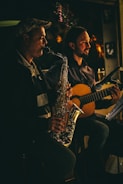 Two musicians are performing in a dimly lit setting. One person is playing a saxophone, and the other is playing a guitar. Both appear focused and engaged in their performance. The lighting casts warm, soft highlights, creating a cozy and intimate atmosphere.