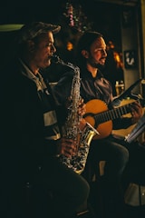 Two musicians are performing in a dimly lit setting. One person is playing a saxophone, and the other is playing a guitar. Both appear focused and engaged in their performance. The lighting casts warm, soft highlights, creating a cozy and intimate atmosphere.