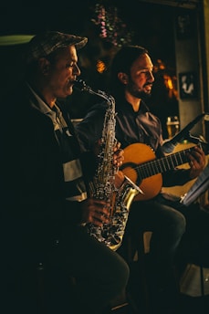 Two musicians are performing in a dimly lit setting. One person is playing a saxophone, and the other is playing a guitar. Both appear focused and engaged in their performance. The lighting casts warm, soft highlights, creating a cozy and intimate atmosphere.