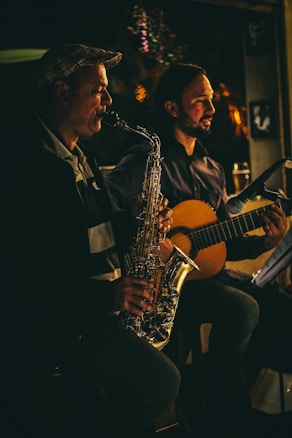 Two musicians are performing in a dimly lit setting. One person is playing a saxophone, and the other is playing a guitar. Both appear focused and engaged in their performance. The lighting casts warm, soft highlights, creating a cozy and intimate atmosphere.