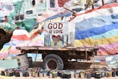 A colorful, mural-painted structure with the words 'God is Love' prominently displayed on a central sign. Below the sign is a collection of paint cans arranged in a semi-circle. The background is vibrant with various hues of paints in abstract patterns.