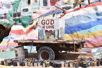 A colorful, mural-painted structure with the words 'God is Love' prominently displayed on a central sign. Below the sign is a collection of paint cans arranged in a semi-circle. The background is vibrant with various hues of paints in abstract patterns.