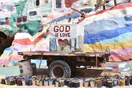 A colorful, mural-painted structure with the words 'God is Love' prominently displayed on a central sign. Below the sign is a collection of paint cans arranged in a semi-circle. The background is vibrant with various hues of paints in abstract patterns.