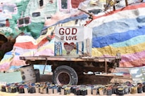 A colorful, mural-painted structure with the words 'God is Love' prominently displayed on a central sign. Below the sign is a collection of paint cans arranged in a semi-circle. The background is vibrant with various hues of paints in abstract patterns.