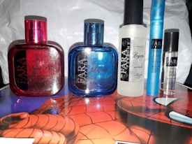 A collection of six perfume bottles placed on a surface decorated with a Spider-Man design. The bottles are of different shapes and sizes, with various branding labels. Two prominent bottles are colored red and blue, while others are white, light blue, and transparent black. The lighting creates reflections and highlights on the bottles and labels.