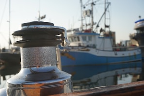 A close-up of a metal winch on a boat with a slightly weathered surface, set against the background of a harbor. Several fishing boats, identifiable by their masts and rigging, are docked nearby. The water reflects portions of the boats and docks, creating a serene and calm setting.