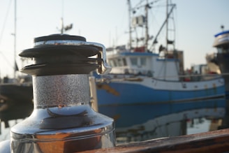 A close-up of a metal winch on a boat with a slightly weathered surface, set against the background of a harbor. Several fishing boats, identifiable by their masts and rigging, are docked nearby. The water reflects portions of the boats and docks, creating a serene and calm setting.