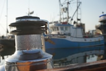 A close-up of a metal winch on a boat with a slightly weathered surface, set against the background of a harbor. Several fishing boats, identifiable by their masts and rigging, are docked nearby. The water reflects portions of the boats and docks, creating a serene and calm setting.