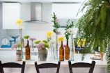 A modern kitchen features a white countertop adorned with various flower arrangements in glass and ceramic vases, alongside brown glass bottles. The background displays white cabinetry, a stainless steel range hood, and a turquoise weighing scale. A lush fern partly obscures the right side of the image, adding greenery to the scene.