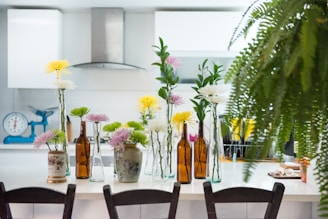 A modern kitchen features a white countertop adorned with various flower arrangements in glass and ceramic vases, alongside brown glass bottles. The background displays white cabinetry, a stainless steel range hood, and a turquoise weighing scale. A lush fern partly obscures the right side of the image, adding greenery to the scene.