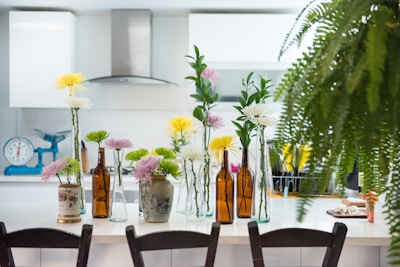 A modern kitchen features a white countertop adorned with various flower arrangements in glass and ceramic vases, alongside brown glass bottles. The background displays white cabinetry, a stainless steel range hood, and a turquoise weighing scale. A lush fern partly obscures the right side of the image, adding greenery to the scene.