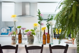 A modern kitchen features a white countertop adorned with various flower arrangements in glass and ceramic vases, alongside brown glass bottles. The background displays white cabinetry, a stainless steel range hood, and a turquoise weighing scale. A lush fern partly obscures the right side of the image, adding greenery to the scene.