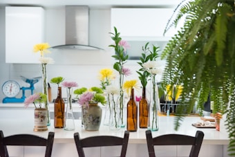 A modern kitchen features a white countertop adorned with various flower arrangements in glass and ceramic vases, alongside brown glass bottles. The background displays white cabinetry, a stainless steel range hood, and a turquoise weighing scale. A lush fern partly obscures the right side of the image, adding greenery to the scene.