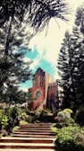 A rustic brick church with a prominent circular window and a cross atop, surrounded by lush green trees and vibrant flowering plants. The church is approached by a staircase lined with well-maintained foliage.