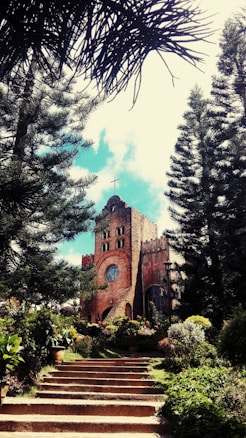 A rustic brick church with a prominent circular window and a cross atop, surrounded by lush green trees and vibrant flowering plants. The church is approached by a staircase lined with well-maintained foliage.