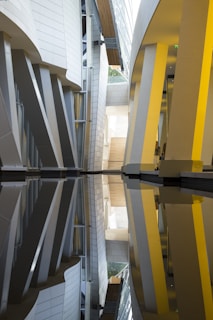 An architectural interior with modern design elements, featuring vertical and slanted pillars alongside reflective surfaces that create symmetrical patterns. Light filters through the spaces between structures, highlighting the interplay of curves and angles.