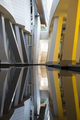An architectural interior with modern design elements, featuring vertical and slanted pillars alongside reflective surfaces that create symmetrical patterns. Light filters through the spaces between structures, highlighting the interplay of curves and angles.