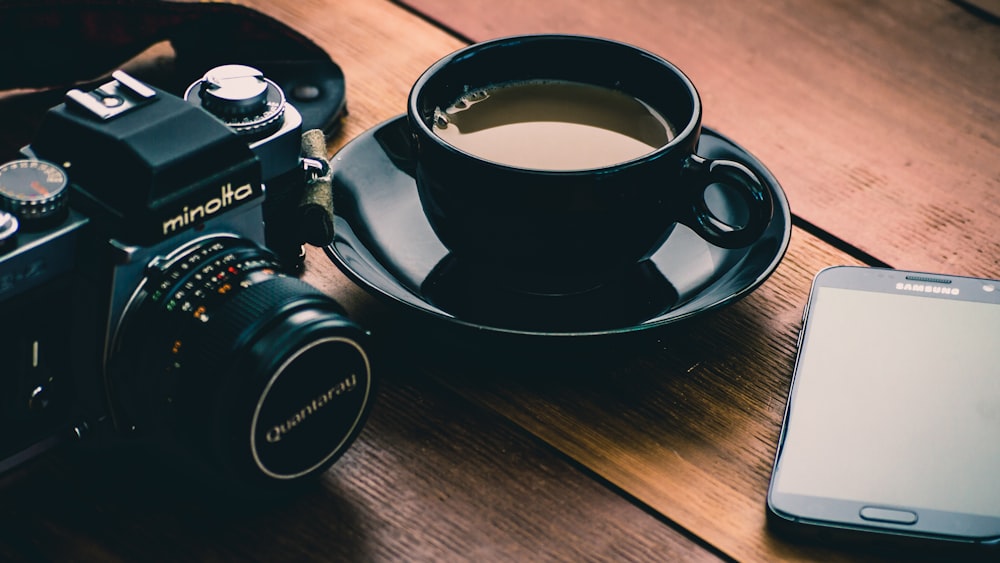 câmera Minolta preta ao lado de xícara de cerâmica preta cheia de café
