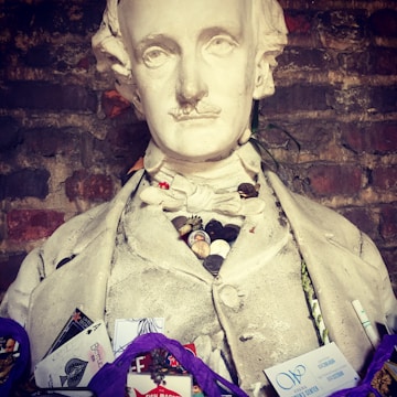 A bust of a man wearing a period outfit adorned with various small items including buttons, cards, and tokens. The bust is set against a brick wall background and has an aged, somewhat solemn appearance.