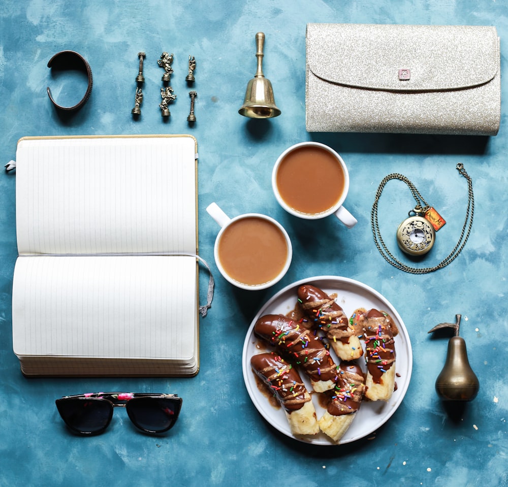 flat lay photography of opened book, coffee mugs, handbag, and sunglasses