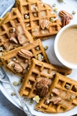 Golden-brown waffles are topped with whole pecans and small white flowers. They are arranged on a white plate with a small bowl of creamy sauce next to them. A silver fork with intricate detailing is also placed on the plate.