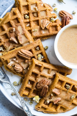 Golden-brown waffles are topped with whole pecans and small white flowers. They are arranged on a white plate with a small bowl of creamy sauce next to them. A silver fork with intricate detailing is also placed on the plate.