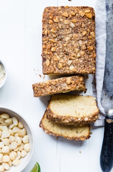 A loaf of bread topped with crushed nuts is partially sliced, with a knife resting on a cloth nearby. Beside the loaf, there is a bowl of whole macadamia nuts and a wedge of lime. The bread has a light, moist texture and is displayed on a white wooden surface.