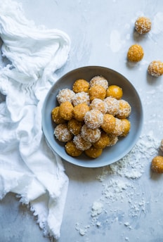 A bowl filled with round yellow-orange sweets, some coated in a white substance presumably coconut. The bowl is placed on a light gray surface, surrounded by loose coconut flakes and a white, textured cloth. Additional sweets are scattered around the table.