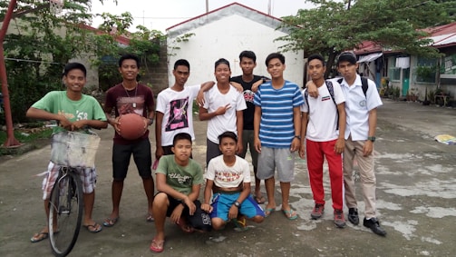 A group of young people are gathered outside, some standing and others squatting. They are casually dressed and appear relaxed and happy. One person has a bicycle, and another holds a basketball. In the background, there are houses and trees.