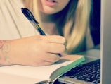 A person is writing in a notebook with a pen while using a laptop. The focus is on the hand and the writing action. A part of the person's face is visible, showcasing concentration.